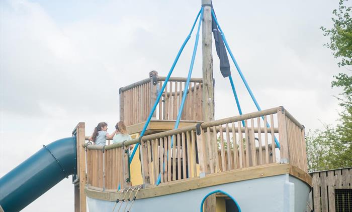 A wooden pirate ship rises from the play area, complete with lookout deck, rope bridge and portholes. Surrounded by green hills, it invites little buc